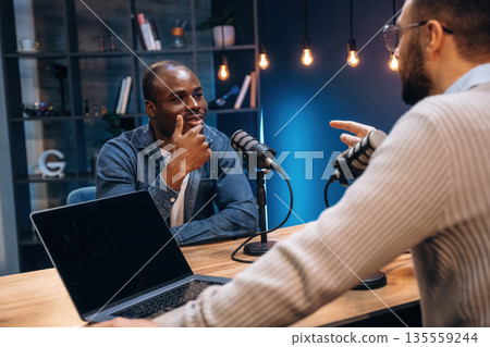 Sitting by the table. Two radio presenters recording an audio broadcast in a studio Sitting by the table. Two radio presenters recording an audio broadcast in a studio 135559244
