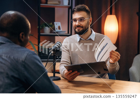 Sitting by the table. Two radio presenters recording an audio broadcast in a studio 135559247
