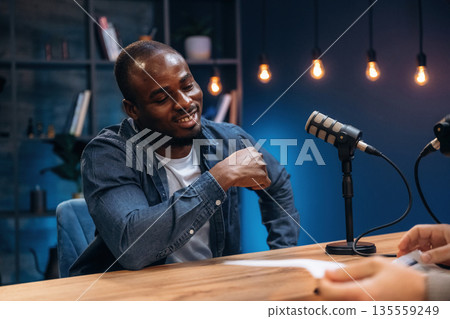Sitting by the table. Two radio presenters recording an audio broadcast in a studio Sitting by the table. Two radio presenters recording an audio broadcast in a studio 135559249