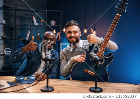 Posing, holding acoustic guitar. Two radio presenters recording an audio broadcast in a studio Posing, holding acoustic guitar. Two radio presenters recording an audio broadcast in a studio 135559337