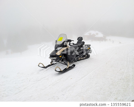 Snowmobile parked on foggy ski area near mountain lodge. Winter transport, resort logistics, and cold weather mobility through technology and alpine service operations. 135559621