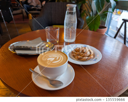 Coffee cup and pastry on wooden table in cozy cafe. Slow morning ritual, relaxation, and mindful break through warm drinks and quiet atmosphere. 135559666
