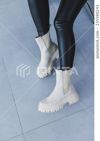 A young beautiful woman is sitting in a chair, wearing white leather boots from a new collection of shoes for spring. A young beautiful woman is sitting in a chair, wearing white leather boots from a new collection of shoes for spring. 135560243