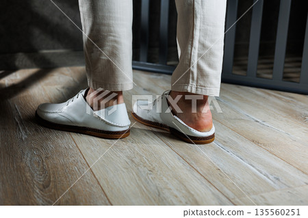 Male feet close-up in white casual shoes. Fashionable young man standing in leather stylish white moccasins in trousers. Seasonal summer men's shoes. Casual street style. 135560251