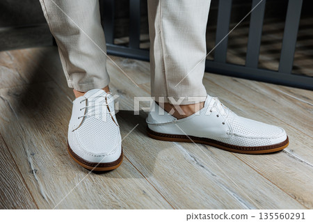 Male feet close-up in white casual shoes. Fashionable young man standing in leather stylish white moccasins in trousers. Seasonal summer men's shoes. Casual street style. Male feet close-up in white casual shoes. Fashionable young man standing in leather stylish white moccasins in trousers. Seasonal summer men's shoes. Casual street style. 135560291