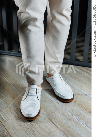 Male feet close-up in white casual shoes. Fashionable young man standing in leather stylish white moccasins in trousers. Seasonal summer men's shoes. Casual street style. Male feet close-up in white casual shoes. Fashionable young man standing in leather stylish white moccasins in trousers. Seasonal summer men's shoes. Casual street style. 135560306
