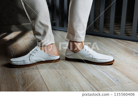 Male feet close-up in white casual shoes. Fashionable young man standing in leather stylish white moccasins in trousers. Seasonal summer men's shoes. Casual street style. Male feet close-up in white casual shoes. Fashionable young man standing in leather stylish white moccasins in trousers. Seasonal summer men's shoes. Casual street style. 135560406