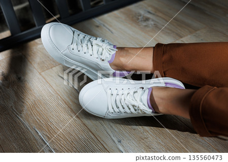 Comfortable female legs in white sneakers with flat soles. Close-up of female legs in leather sneakers. Women's summer sneakers. Collection of women's leather shoes Comfortable female legs in white sneakers with flat soles. Close-up of female legs in leather sneakers. Women's summer sneakers. Collection of women's leather shoes 135560473