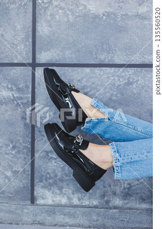 A young woman with slender beautiful legs in fashionable black leather shoes sits on a chair in the studio. Women's stylish loafers, close-up. Spring shoe collection A young woman with slender beautiful legs in fashionable black leather shoes sits on a chair in the studio. Women's stylish loafers, close-up. Spring shoe collection 135560520