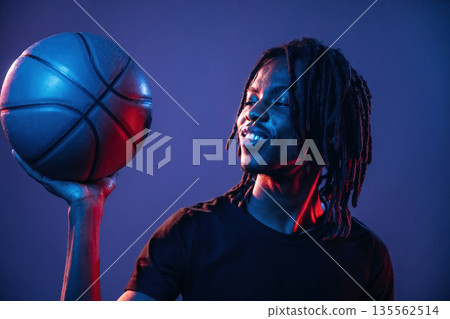 Dark room, basketball ball. African American guy is studio with neon lighting Dark room, basketball ball. African American guy is studio with neon lighting 135562514