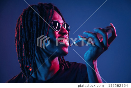 Stylish sunglasses, talking by phone. African American guy is studio with neon lighting Stylish sunglasses, talking by phone. African American guy is studio with neon lighting 135562564