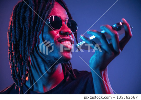 Stylish sunglasses, talking by phone. African American guy is studio with neon lighting Stylish sunglasses, talking by phone. African American guy is studio with neon lighting 135562567