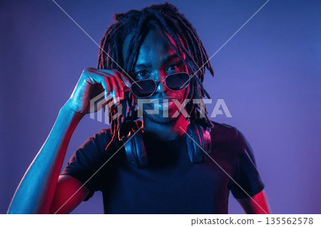 Touching sunglasses, front view. African American guy is studio with neon lighting Touching sunglasses, front view. African American guy is studio with neon lighting 135562578