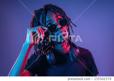 Touching sunglasses, front view. African American guy is studio with neon lighting 135562579