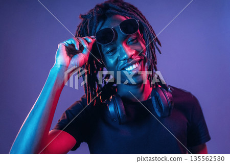 Touching sunglasses, front view. African American guy is studio with neon lighting 135562580
