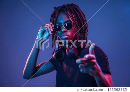 Pointing by finger, in sunglasses. African American guy is studio with neon lighting 135562585