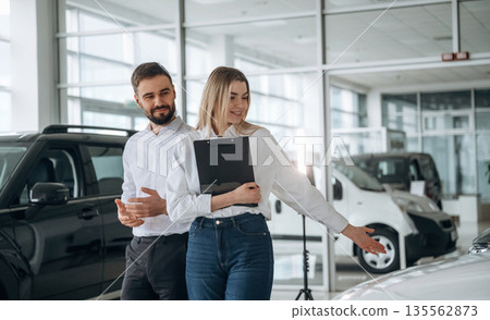 Black notepad in hands. Man and woman are in the car dealership Black notepad in hands. Man and woman are in the car dealership 135562873