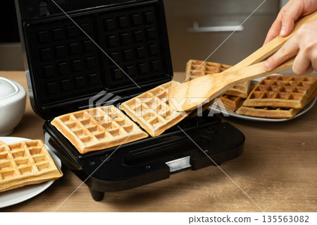 Taking Out Hot Golden-Brown Waffles from an Electric Maker in a Home Kitchen. Taking Out Hot Golden-Brown Waffles from an Electric Maker in a Home Kitchen. 135563082