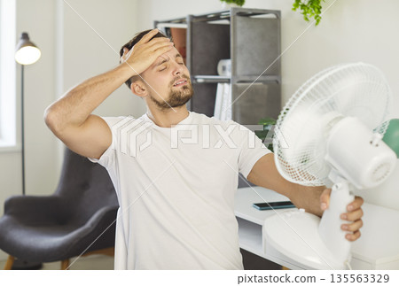 Man is standing indoors, feeling too hot and wiping forehead with hand, holding fan blowing air Man is standing indoors, feeling too hot and wiping forehead with hand, holding fan blowing air 135563329