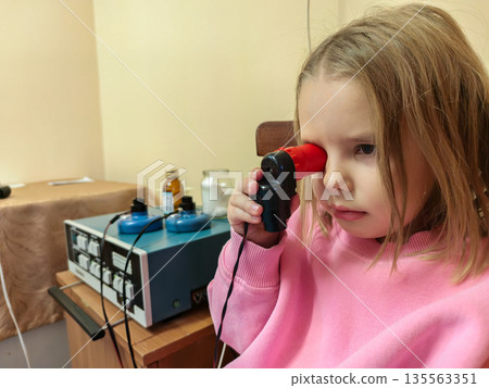 A girl is undergoing magnetic eye therapy at a modern clinic. During physiotherapy, the child receives targeted treatment using specialized equipment for vision rehabilitation and medical care. A girl is undergoing magnetic eye therapy at a modern clinic. During physiotherapy, the child receives targeted treatment using specialized equipment for vision rehabilitation and medical care. 135563351