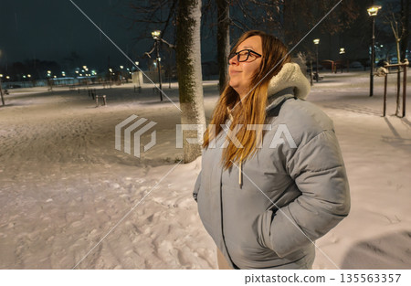 A woman stands in a quiet, snow-covered park on a cold winter night, eyes closed, enjoying the peaceful atmosphere under the streetlights. A woman strolls through a winter park in the evening. A woman stands in a quiet, snow-covered park on a cold winter night, eyes closed, enjoying the peaceful atmosphere under the streetlights. A woman strolls through a winter park in the evening. 135563357