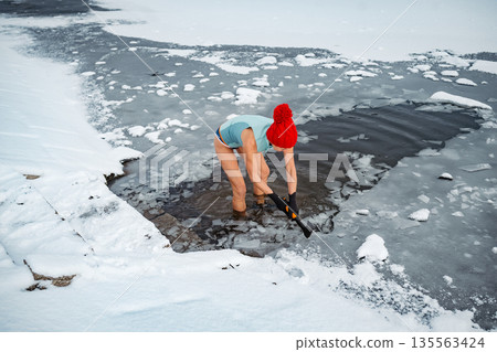 Senior woman breaking ice for winter swimming on frozen lake. 135563424