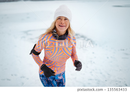 Active elderly woman jogging in cold snowy weather. 135563433
