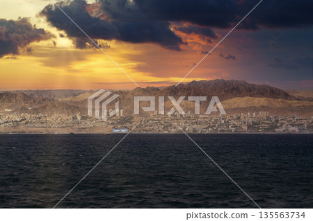 View of a coastal Eilat city at sunset with mountains in the background and dark clouds above sea 135563734