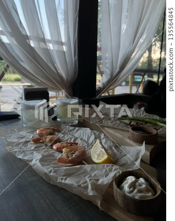 A delicious serving of shrimp garnished with lemon awaits on a rustic wooden table. Two refreshing drinks accompany the seafood, perfect for sharing at a meal 135564845