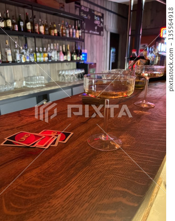 Two elegant cocktail glasses rest on a polished wooden bar, with UNO cards nearby. The vibrant backdrop suggests an inviting social setting perfect for games 135564918
