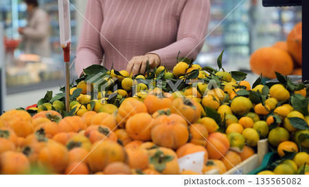 Young female choosing fresh tangerines at grocery near a persimmon display, healthy eating and organic lifestyle Young female choosing fresh tangerines at grocery near a persimmon display, healthy eating and organic lifestyle 135565082