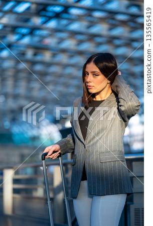Businesswoman dressed in a pinstripe blazer and black top carrying a travel suitcase, walking through an modern airport terminal structure on a sunny day 135565419