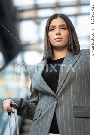 Young professional woman commuting or embarking on a business trip, standing with her luggage handle in what appears to be an airport or train station environment Young professional woman commuting or embarking on a business trip, standing with her luggage handle in what appears to be an airport or train station environment 135565423