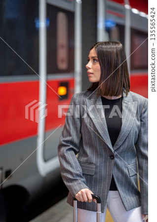 Young professional woman in a pinstripe blazer stands on a train platform with a suitcase, waiting confidently for a business trip in an urban commuter station Young professional woman in a pinstripe blazer stands on a train platform with a suitcase, waiting confidently for a business trip in an urban commuter station 135565424