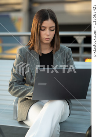 Young businesswoman focusing on her laptop screen, working efficiently outdoors in an urban setting, embracing remote work and digital nomad lifestyle Young businesswoman focusing on her laptop screen, working efficiently outdoors in an urban setting, embracing remote work and digital nomad lifestyle 135565425
