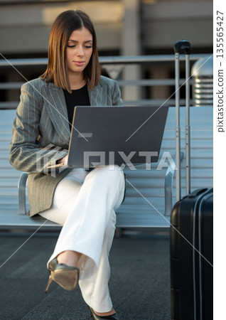 Businesswoman sitting on a bench at an airport or train station, focusing on her laptop with luggage beside her, embodying concepts of global business, remote work, and professional travel Businesswoman sitting on a bench at an airport or train station, focusing on her laptop with luggage beside her, embodying concepts of global business, remote work, and professional travel 135565427