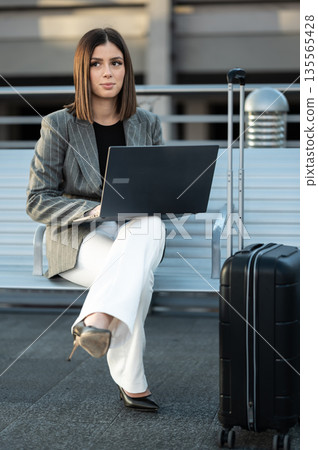 Young businesswoman sitting on a bench, typing on her laptop while her black suitcase waits beside her, embodying remote work and travel convenience in a professional setting Young businesswoman sitting on a bench, typing on her laptop while her black suitcase waits beside her, embodying remote work and travel convenience in a professional setting 135565428