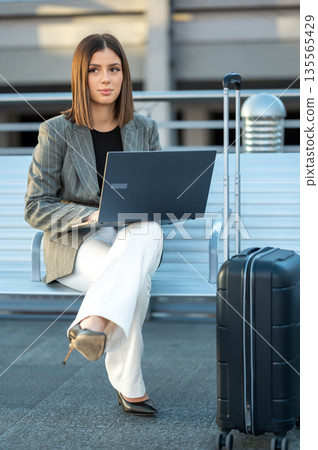 Young businesswoman sitting outside an airport terminal working remotely on her laptop while waiting for her flight, showcasing global connectivity and modern business mobility 135565429