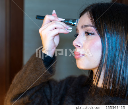 Young woman applying liquid foundation under her eye with a specialized brush, creating a clean beauty look and emphasizing cosmetic application for skin care Young woman applying liquid foundation under her eye with a specialized brush, creating a clean beauty look and emphasizing cosmetic application for skin care 135565435
