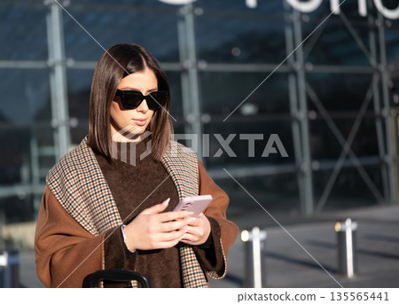 Stylish woman wearing sunglasses and a patterned scarf, standing outside a modern airport building, checking her smartphone while preparing for departure, embodying travel and digital connection 135565441