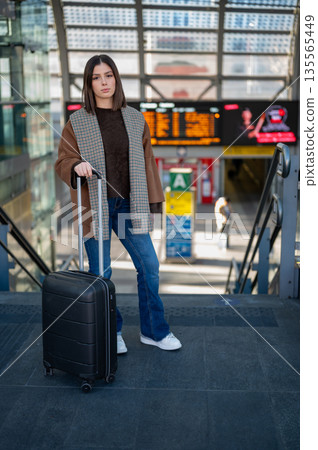 Young woman standing on a platform holding a black suitcase, ready for a journey at a modern train station with a departure board in the background, representing travel and transportation concepts Young woman standing on a platform holding a black suitcase, ready for a journey at a modern train station with a departure board in the background, representing travel and transportation concepts 135565449