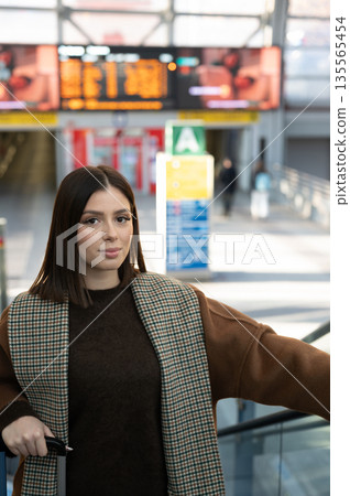 Young woman standing on an escalator holding a luggage handle, preparing for a professional or personal journey inside a modern transportation hub with a departure board in the background 135565454