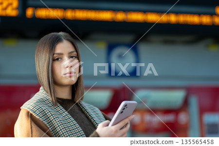 Young woman standing on a train station platform, holding her mobile phone and waiting for public transport, with a blurred information board in the background Young woman standing on a train station platform, holding her mobile phone and waiting for public transport, with a blurred information board in the background 135565458