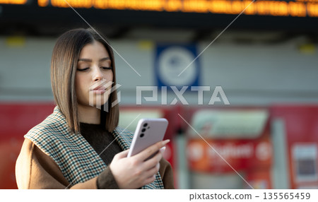 Young woman with brown hair in a coat checking her smartphone at a busy train station, waiting by the departure board and staying connected while commuting or traveling Young woman with brown hair in a coat checking her smartphone at a busy train station, waiting by the departure board and staying connected while commuting or traveling 135565459