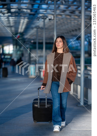 Young woman confidently walking through a modern airport terminal, pulling her black suitcase, representing travel, business trips, and vacation departures 135565463
