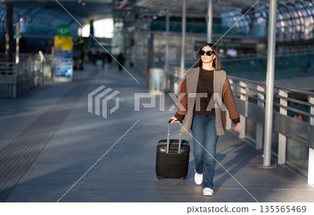 Woman in sunglasses and casual clothes pulls a wheeled suitcase through a bright airport or train station concourse, walking confidently on a solo urban journey between destinations Woman in sunglasses and casual clothes pulls a wheeled suitcase through a bright airport or train station concourse, walking confidently on a solo urban journey between destinations 135565469