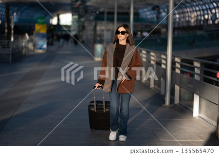 Young female traveler with sunglasses and a brown coat pulling a suitcase, walking through a bright modern airport or train station during transit, representing business trips and leisure concepts Young female traveler with sunglasses and a brown coat pulling a suitcase, walking through a bright modern airport or train station during transit, representing business trips and leisure concepts 135565470
