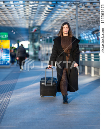 Woman walking confidently through a modern airport or train station terminal, pulling a black wheeled suitcase along the concourse, embodying business travel, holidays, and departure 135565473
