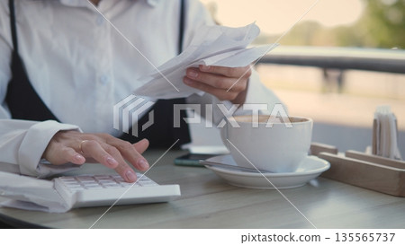 A woman sits at a cafe table, organizing receipts while using a calculator. She enjoys a warm beverage, focused on managing her finances during a sunny afternoon. 135565737