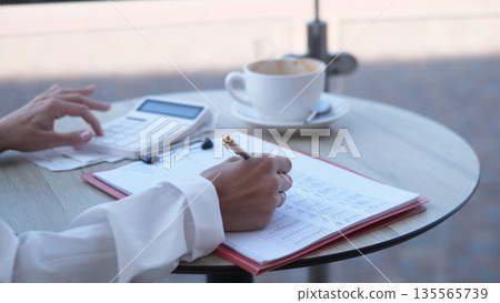 A businesswoman is sitting at a cafe table, engaged in financial calculations. She writes in a notebook with a calculator nearby and coffee in front of her, working on her business plans. 135565739